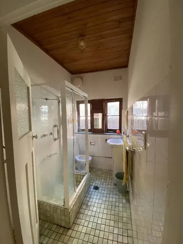 A narrow bathroom with a varnished timber-lined ceiling, a dark-stained window frame and cream walls above white wall tiles.