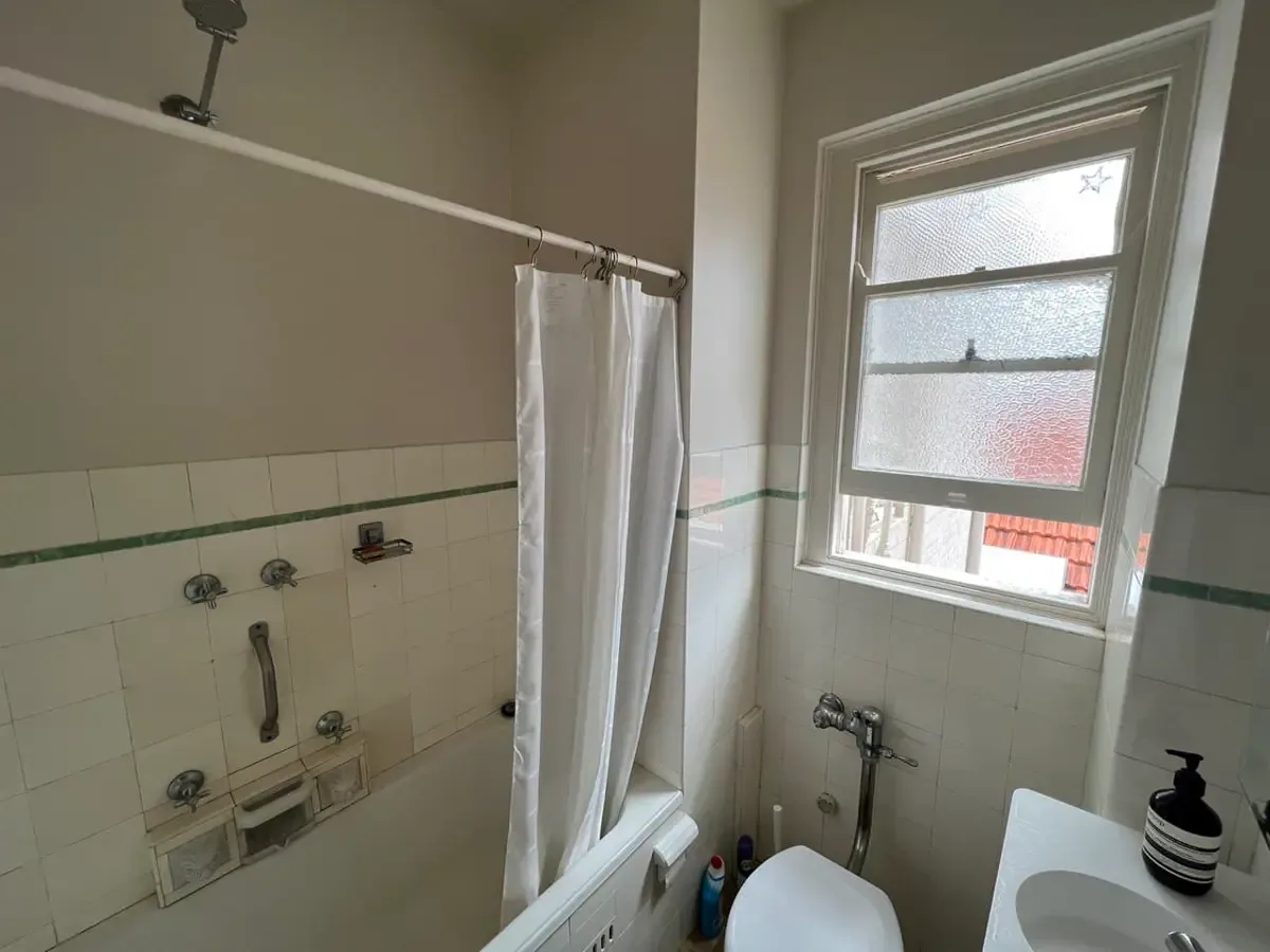 A small bathroom with dull beige walls above cream wall tiles with a green border stripe, and a shower curtain over the bath.