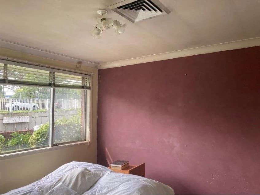A bedroom with a blotchy dark maroon feature wall behind the bed and a discoloured ceiling.