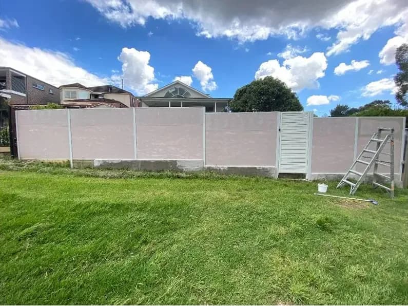 A long backyard boundary fence in a patchy pink-beige, with a white lattice gate and a ladder leaning against the far end.
