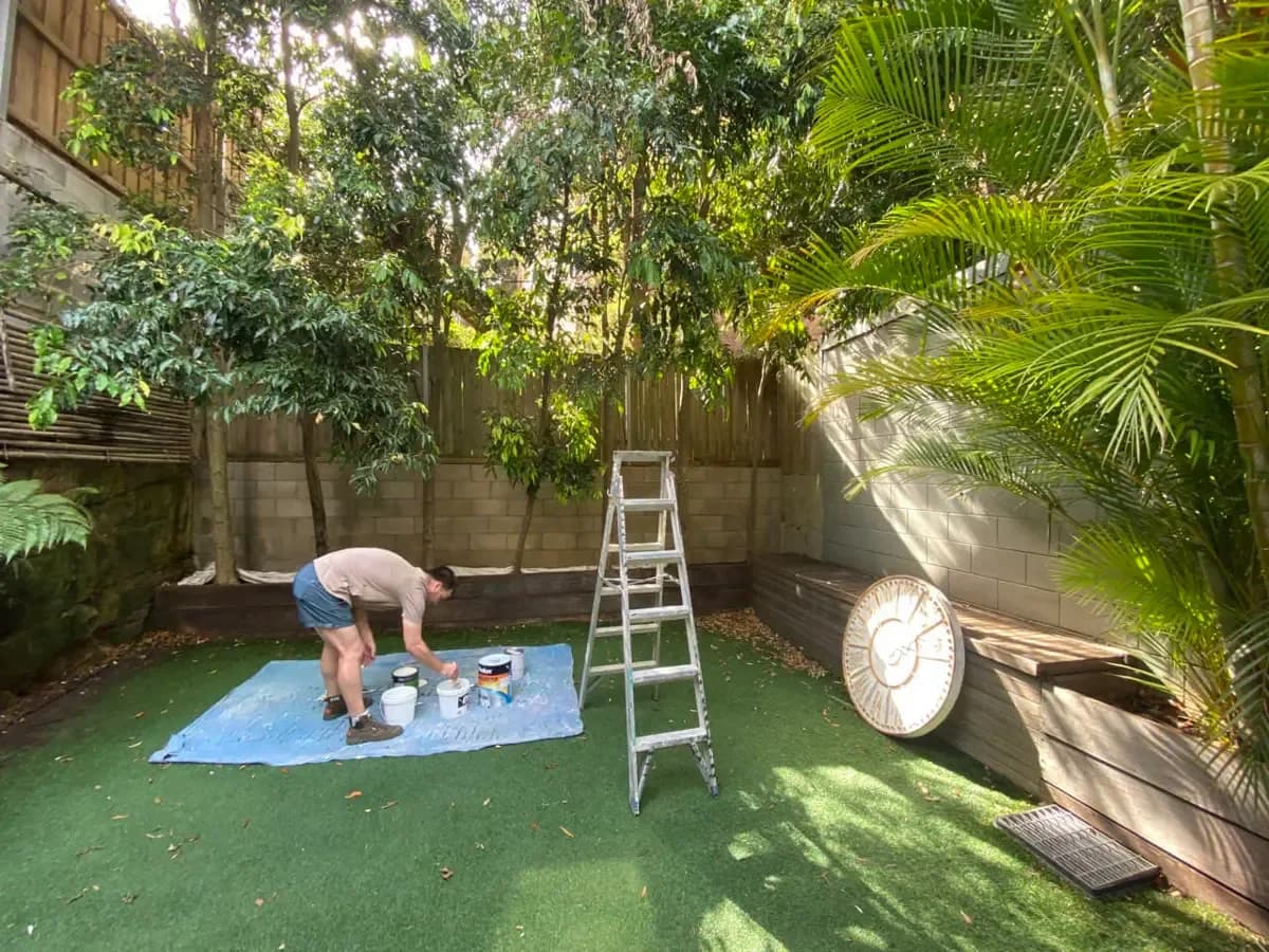 A shaded courtyard with a grey concrete block wall and weathered timber paling fence, and a painter setting out tins on a drop sheet.