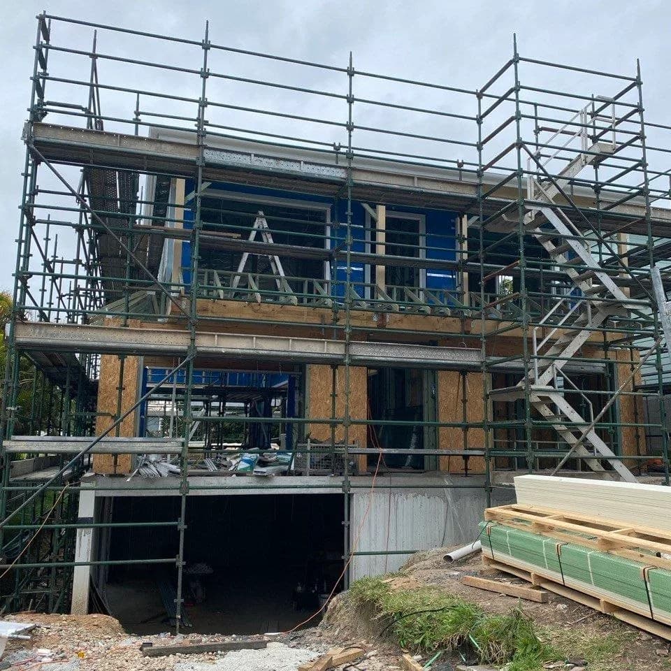 A two-storey weatherboard home mid-build, wrapped in scaffolding with bare framing and blue sarking exposed.