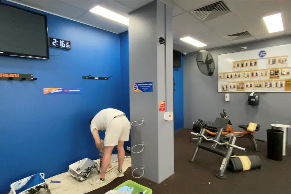 The same gym with the walls finished in a deep blue and the column in grey, a painter cutting in at floor level beside the equipment.