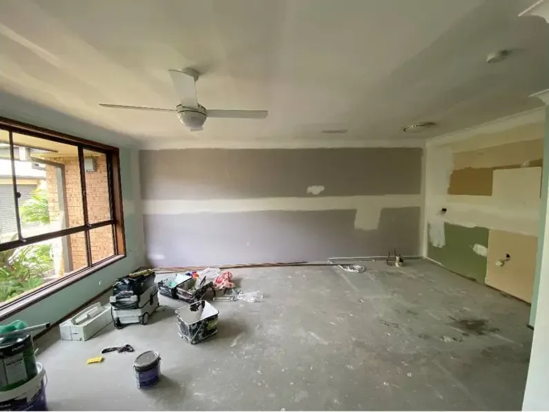 An empty room mid-job with patched plasterboard walls in mismatched grey and green undercoat, and painting gear on the bare floor.