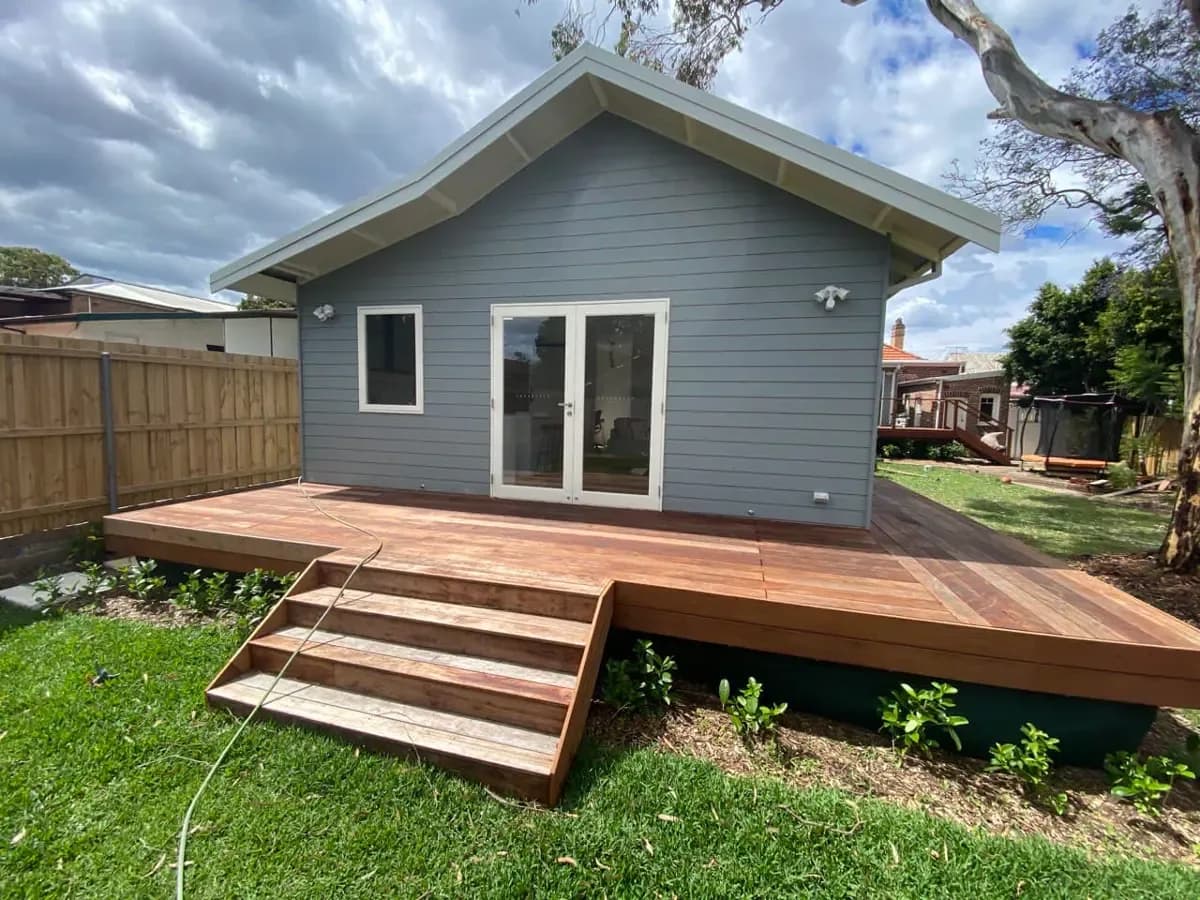 A newly laid timber deck and steps in pale, untreated boards, in front of a grey weatherboard studio with white French doors.
