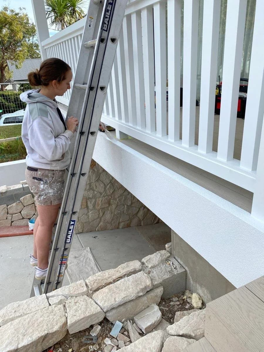 A McGill Painting painter on a ladder, cutting in white trim beneath a verandah balustrade on a weatherboard home.