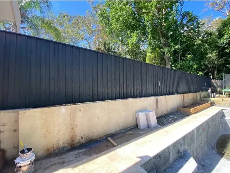 The same fence run coated in a near-black charcoal, standing against the concrete retaining wall and the trees behind it.