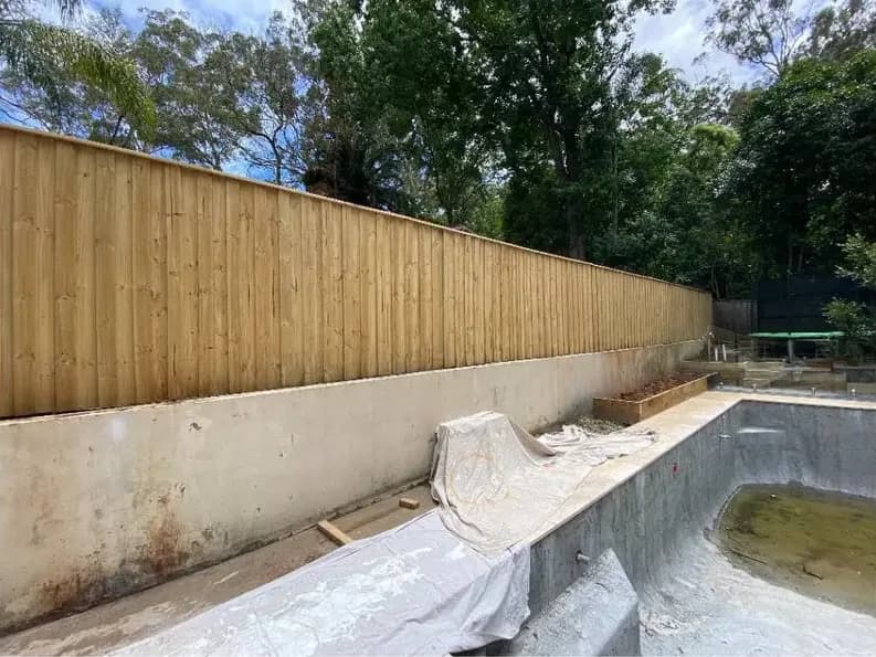 A long timber paling fence in bare pine running above a concrete retaining wall, beside an unfinished pool shell.