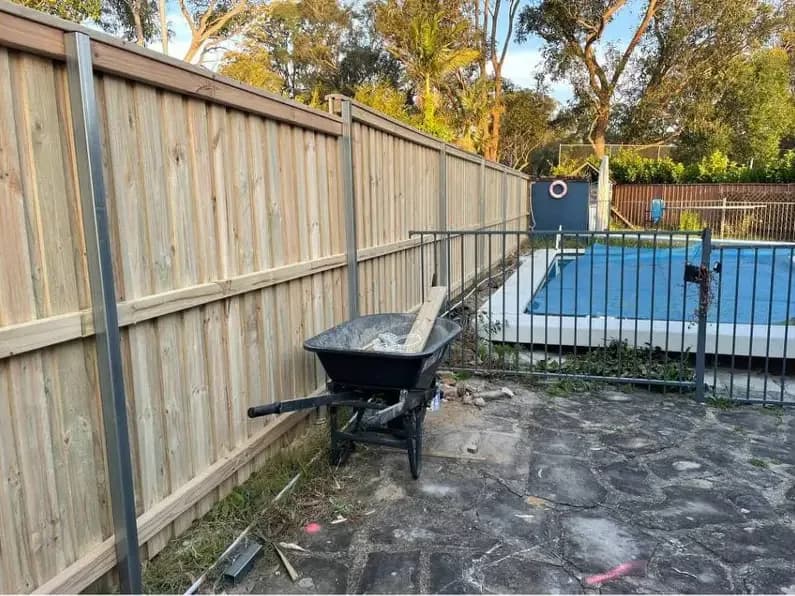 A newly built timber paling fence with bare pine palings and grey steel posts, beside an unfinished pool surround with a wheelbarrow on the concrete.
