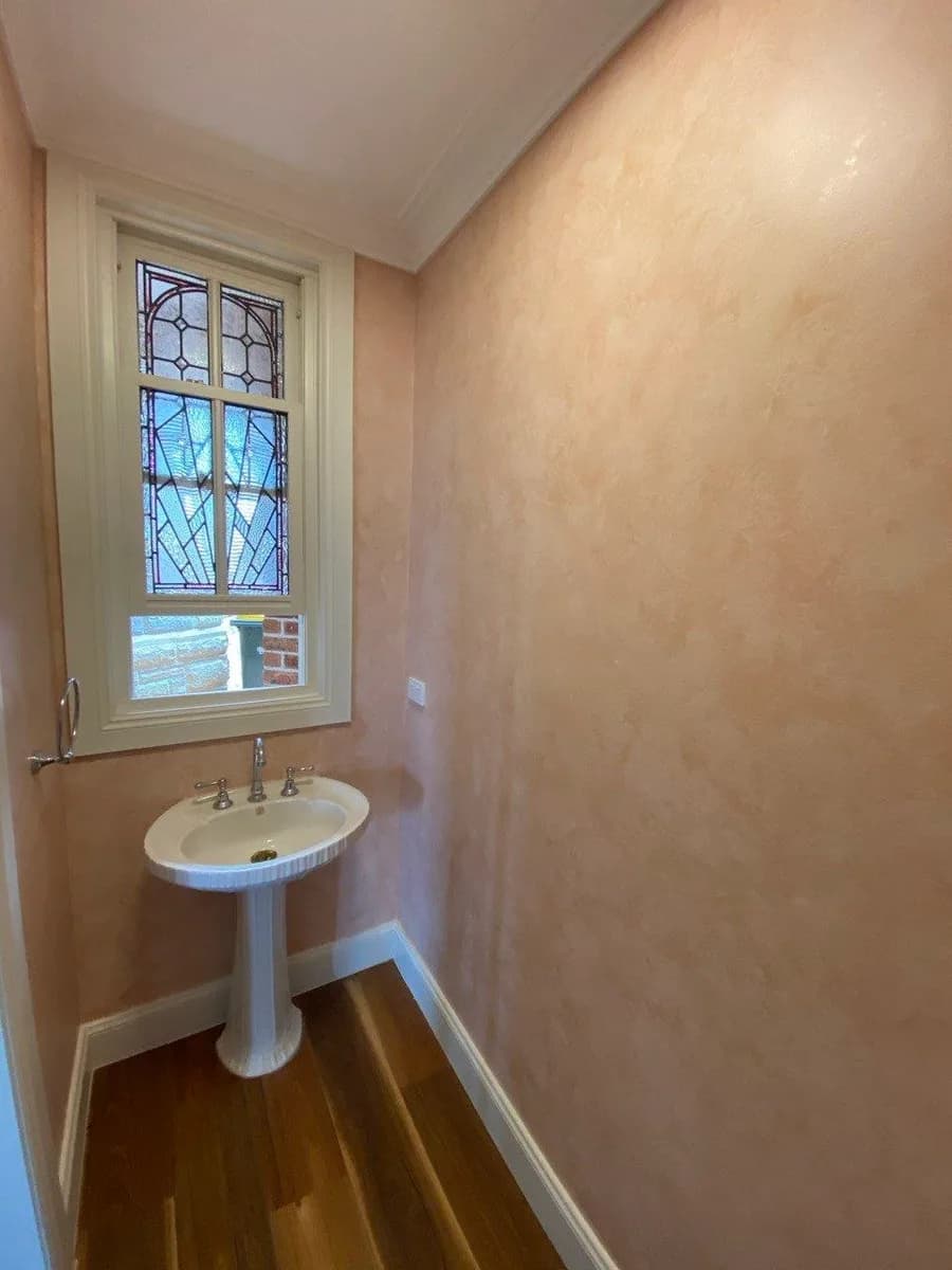 The same powder room finished in a mottled rose French wash — a warm, hand-worked decorative paint effect — with the leadlight window and cornice cut in cleanly.