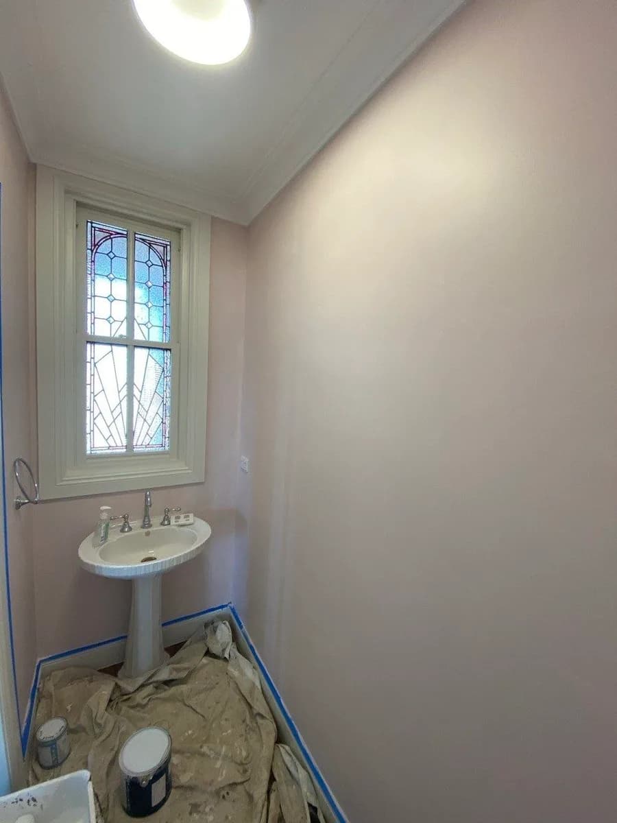 A narrow powder room in a flat, even pale pink, masked up with painter's tape along the skirting and drop sheets down, beside a leadlight window.