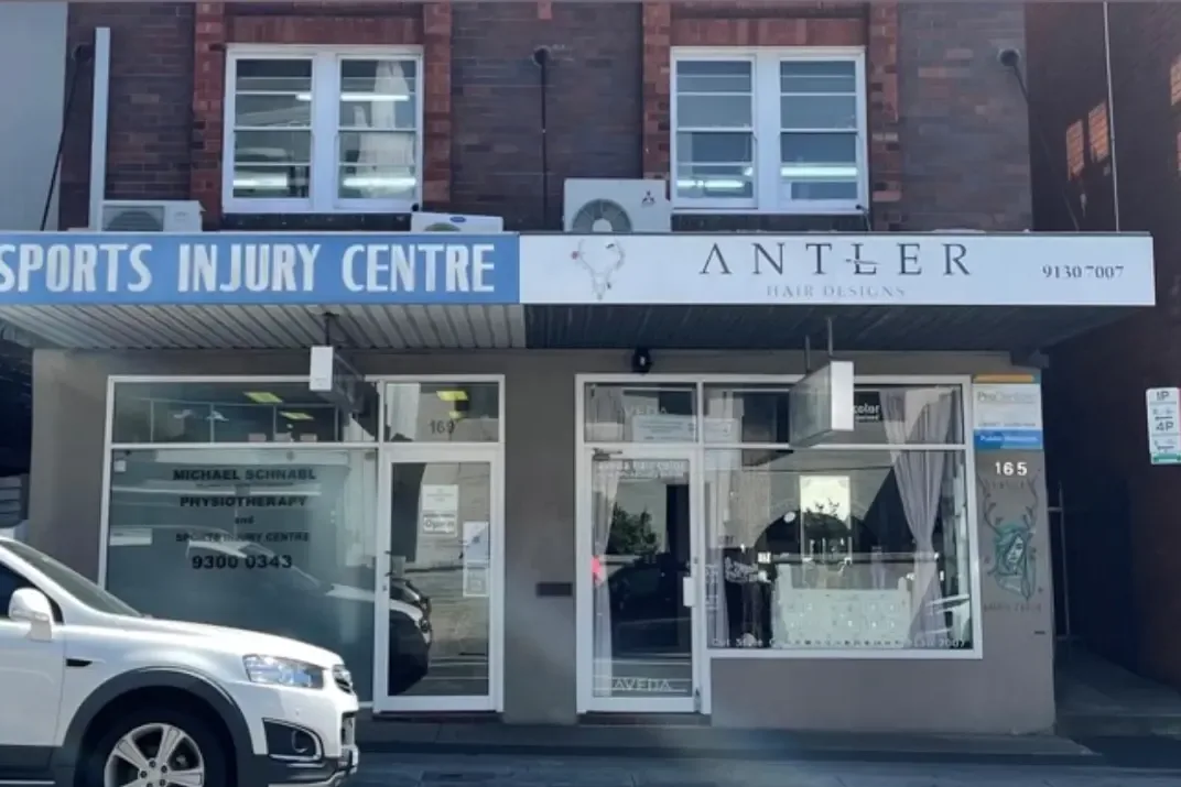 A street-level shopfront strip with tired grey-beige rendered walls and columns beneath a blue and white signage band.